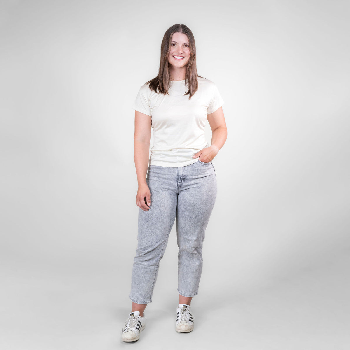 A full body photo of a smiling brown haired woman standing against a white background. She is wearing white sneakers, gray jeans, and a natural white Alpacas of Montana lightweight athletic activewear outerwear breathable moisture wicking antimicrobial soft comfortable exercise short sleeve alpaca women&#39;s performance tee for running sports exercise work out hiking climbing camping biking