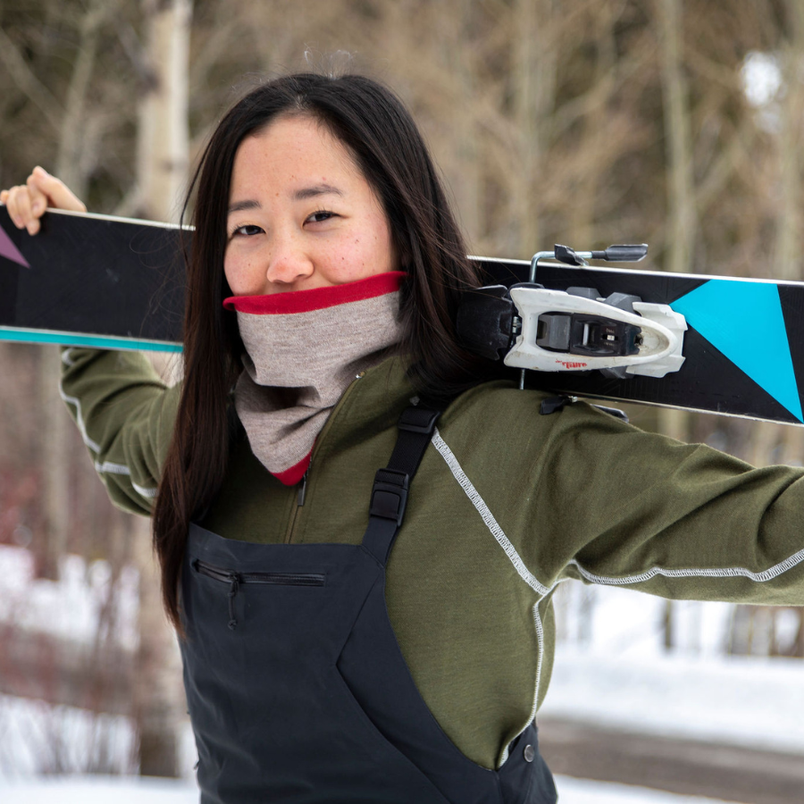 A black-haired woman standing outdoors holding a pair of skis across her shoulders. She is wearing overall snowpants and an olive green Alpacas of Montana women&#39;s base layer top, and a lightweight cozy comfortable moisture wicking warm breathable all seasons thin thermal outerwear skiing hiking climbing outdoors hunting fishing high desert neck gaiter made from scarlet red and light camel brown alpaca.