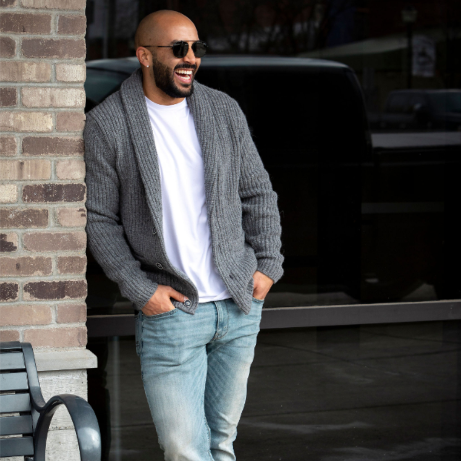 A man leaning against a brick wall casually and laughing. He is wearing sunglasses, light blue jeans, a white shirt, and an Alpacas of Montana cozy comfortable soft moisture wicking formal casual stylish upcycled men&#39;s medium gray button up chunky knit ribbed cardigan.