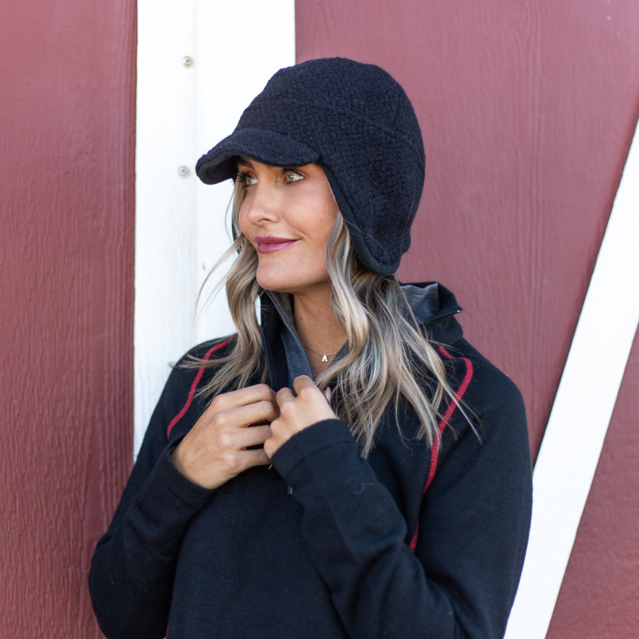 A blonde woman standing in front of a red barn and looking off to her right. She is wearing a black and red Alpacas of Montana women&#39;s mid-layer quarter zip top and a black Alpacas of Montana windstopper extreme warmth hat