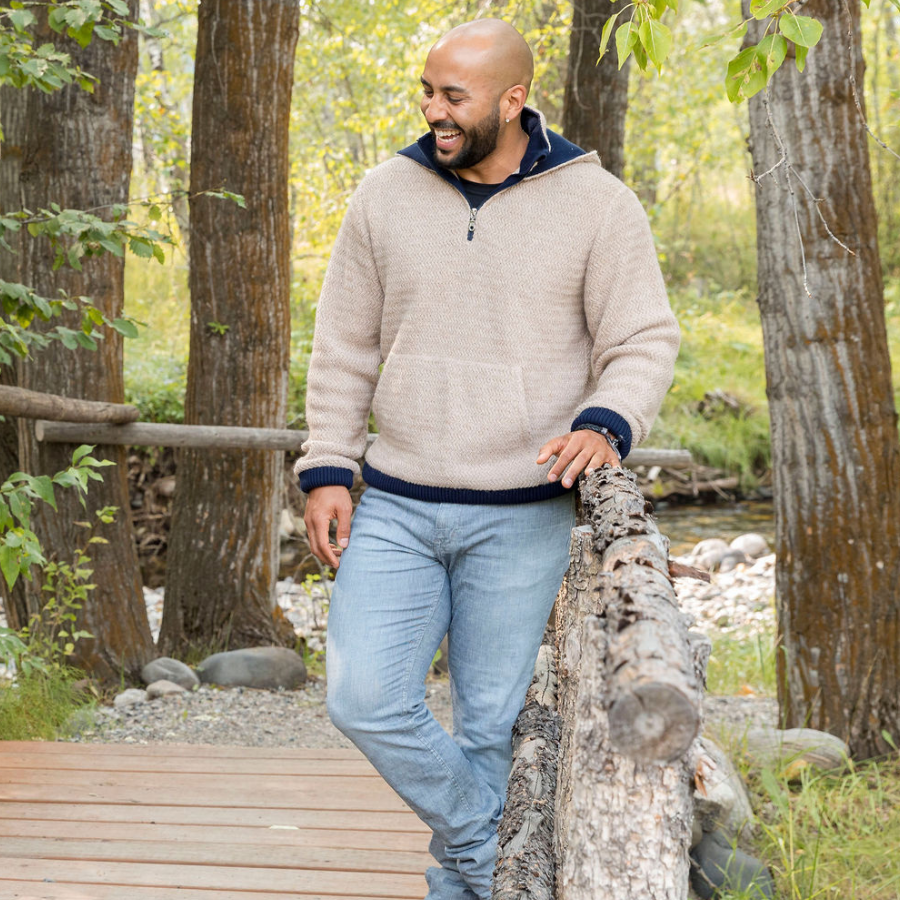 A smiling bald man with a black beard standing on a wooden walkway with wooden railings in a forest. He is wearing blue jeans and a sand cream oatmeal camel color with navy accents Alpacas of Montana thick warm cozy comfortable soft lined heavyweight warmth moisture wicking antimicrobial stylish men&#39;s fashion oversized alpaca wool hoodie.