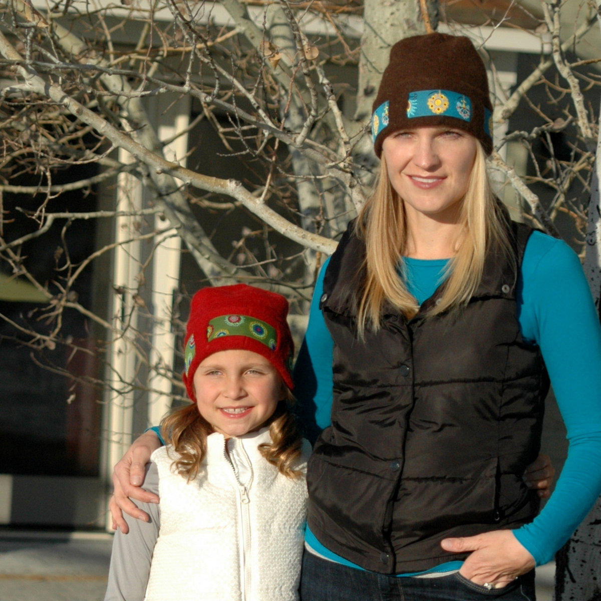 A brown haired girl and a blonde woman standing side by side outdoors. The little girl is wearing a white vest and a scarlet red soft cozy cute warm winter thermal moisture wicking felted alpaca wool beanie hat with a green floral ribbon for kids children. The woman is wearing a cyan long sleeve shirt and black vest with a brown soft cozy cute warm winter thermal moisture wicking felted alpaca wool beanie hat with a teal floral ribbon.