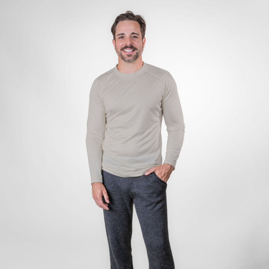 A photo of a smiling man standing against a white background with his hand in his pocket. He is wearing gray pants and a lightweight athletic activewear outerwear breathable moisture wicking antimicrobial soft comfortable exercise long sleeve natural white alpaca men&#39;s performance tee for running sports exercise work out hiking climbing camping hunting skiing biking