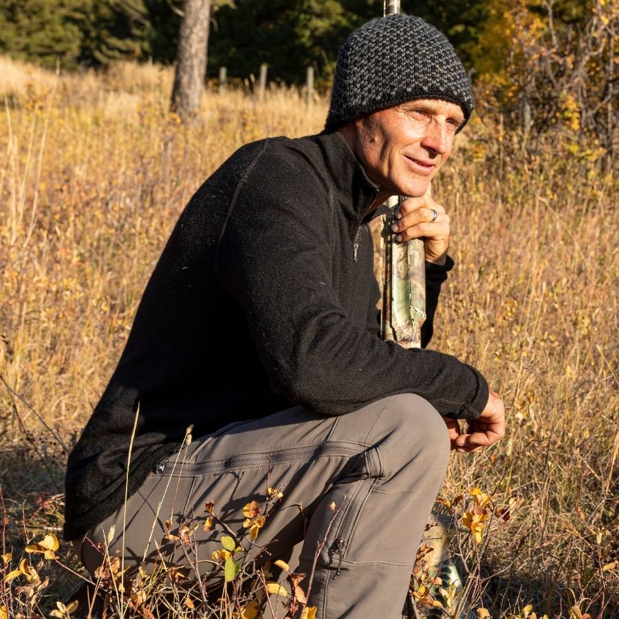 A man kneeling in a golden grassy field looking off into the distance. He is wearing gray pants, a black Alpacas of Montana men&#39;s mid-layer quarter zip top, and a black and gray Alpacas of Montana extremely warm cozy soft windproof comfortable antimicrobial moisture wicking thermal alpaca fleece wool winter fleece lined beanie hat for hiking, skiing, snowshoeing, sledding, outdoors, hunting.