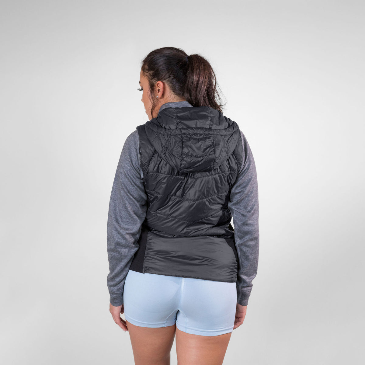 A brown haired woman facing away from the camera in front of a white background. She is wearing light blue athletic shorts, a gray Alpacas of Montana women&#39;s base layer top, and a black Alpacas of Montana lightweight athletic activewear outerwear breathable moisture wicking antimicrobial soft comfortable exercise alpaca women&#39;s stretch lite vest for running sports exercise work out hiking climbing camping biking