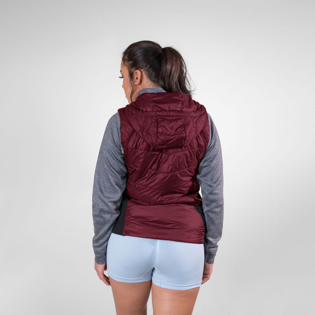 A brown haired woman facing away from the camera in front of a white background. She is wearing light blue athletic shorts, a gray Alpacas of Montana women&#39;s base layer top, and a cranberry red Alpacas of Montana lightweight athletic activewear outerwear breathable moisture wicking antimicrobial soft comfortable exercise alpaca women&#39;s stretch lite vest for running sports exercise work out hiking climbing camping biking