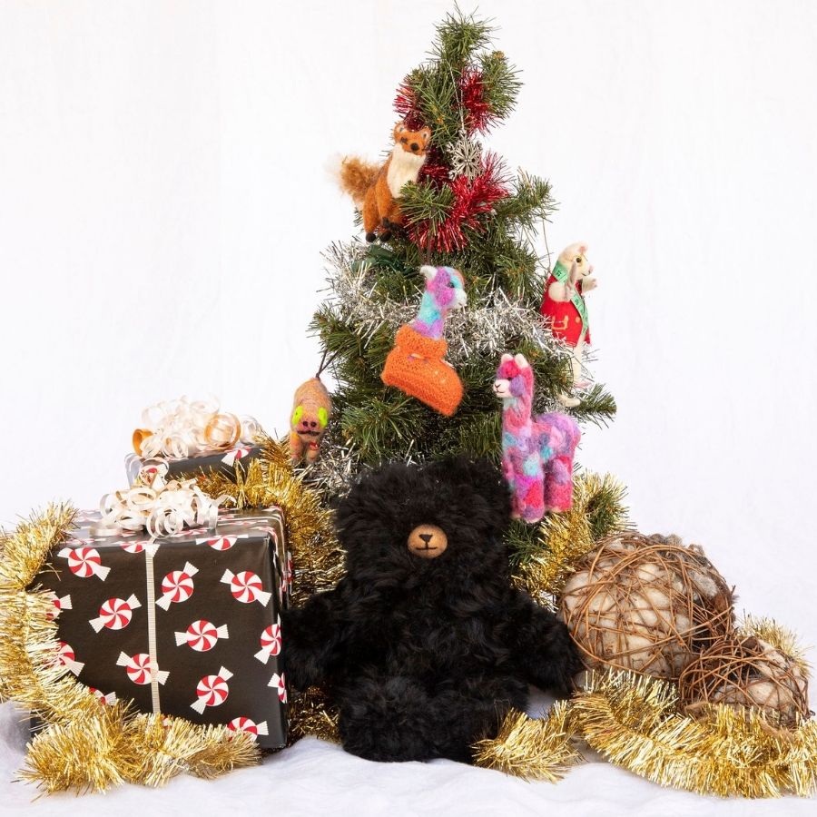 A product photo with a white background of a Christmas tree with Alpacas of Montana figurine ornaments, wrapped presents, tinsel strands, Alpacas of Montana bird balls, and an Alpacas of Montana soft fluffy cozy cute adorable toy silky black luxury royal alpaca teddy bear plushie figurine for gifts birthdays christmas children present