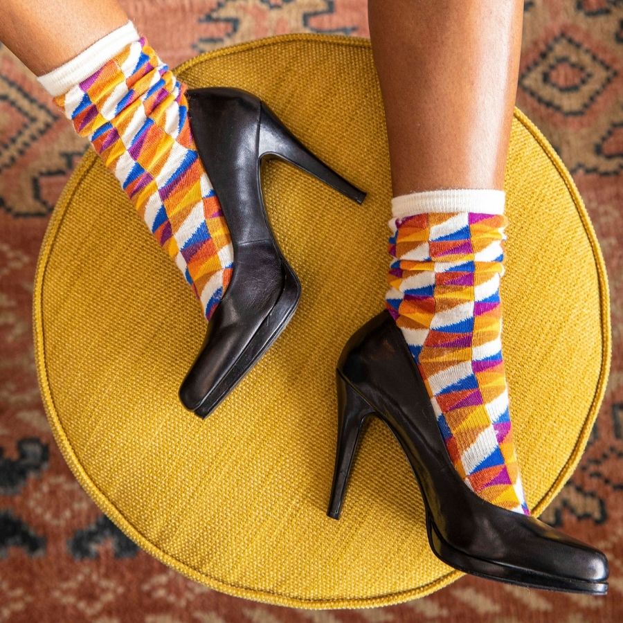A close up of a pair of feet on a woven golden yellow stool wearing black heeled shoes and a pair of Alpacas of Montana white, yellow, gold, orange, purple, and blue triangle pattern casual lounge fashion comfortable soft cozy everyday moisture wicking alpaca wool Swag socks.
