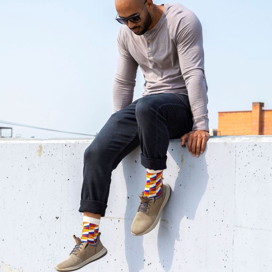 A man sitting on a stone wall looking downward. He is wearing a long sleeve gray shirt, black jeans, tan shoes, and a pair of Alpacas of Montana white, yellow, gold, orange, purple, and blue triangle pattern casual lounge fashion comfortable soft cozy everyday moisture wicking alpaca wool Swag socks.
