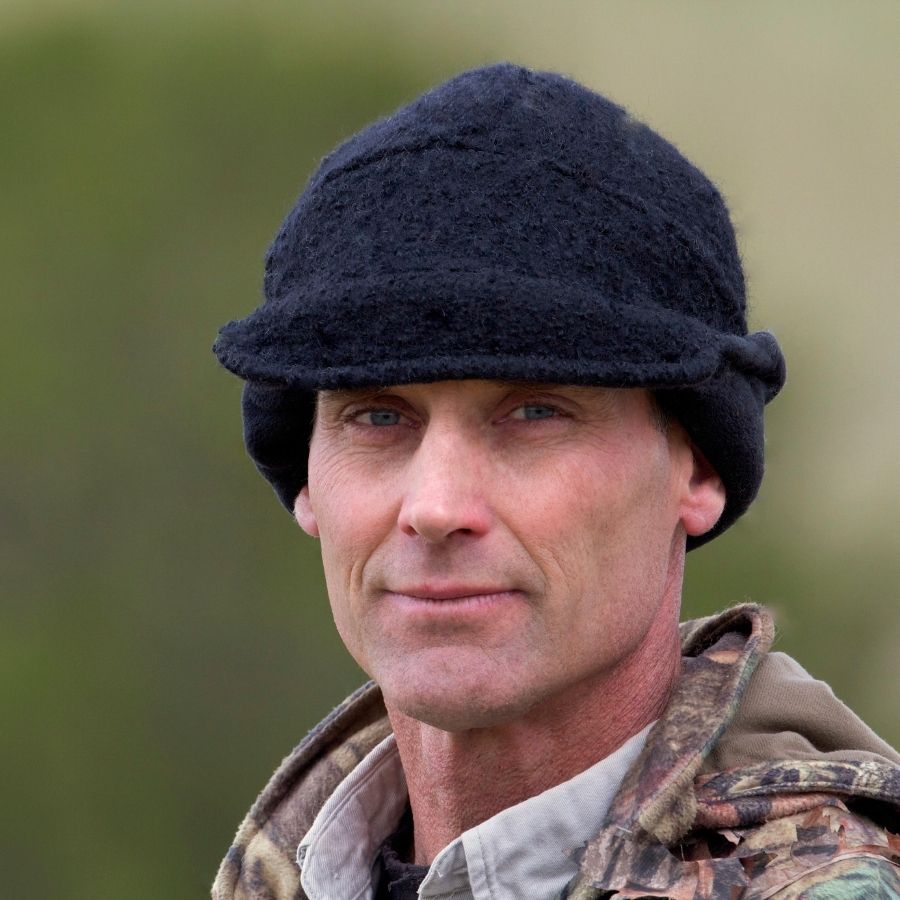 A close up photo of a man looking at the camera wearing camouflage hunting clothing and an extremely warm cozy soft windproof comfortable moisture wicking thermal alpaca fleece wool black windstopper winter hat for hiking, skiing, hunting, fishing, outdoors.