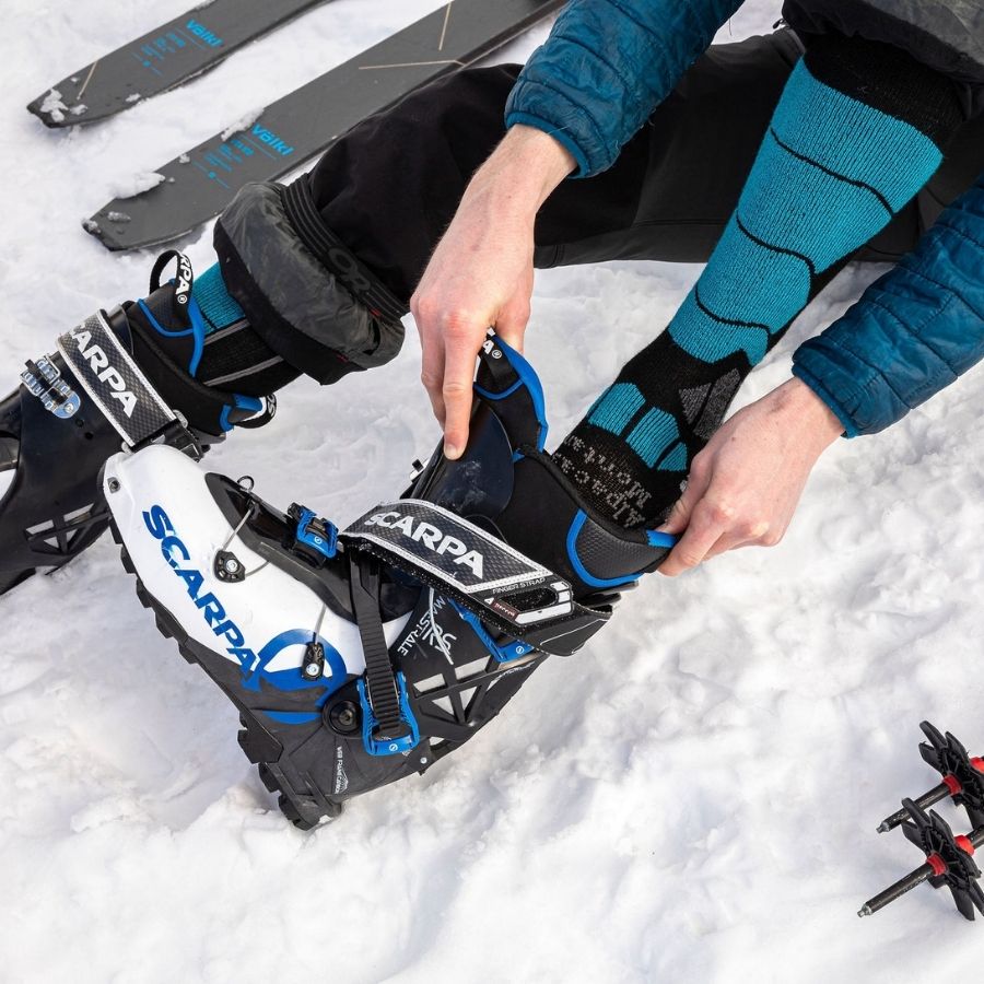 A person sitting in the snow putting their foot into a black, white and blue ski boot. They are wearing a pair of teal blue, gray, and black Alpacas of Montana cozy comfortable soft warm thermal winter freezing temperatures antimicrobial moisture wicking alpaca wool ski and snowboard socks for snowshoeing, skiing, snowboarding, ice fishing, outdoors.