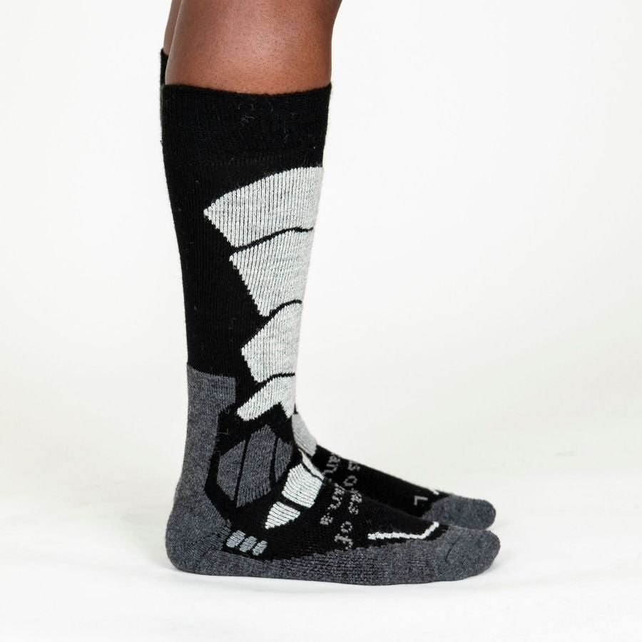 A person&#39;s lower legs standing sideways with a white background wearing light gray, dark grey, and black Alpacas of Montana cozy comfortable soft warm thermal winter freezing temperatures antimicrobial moisture wicking alpaca wool ski and snowboard socks for snowshoeing, skiing, snowboarding, ice fishing, outdoors.