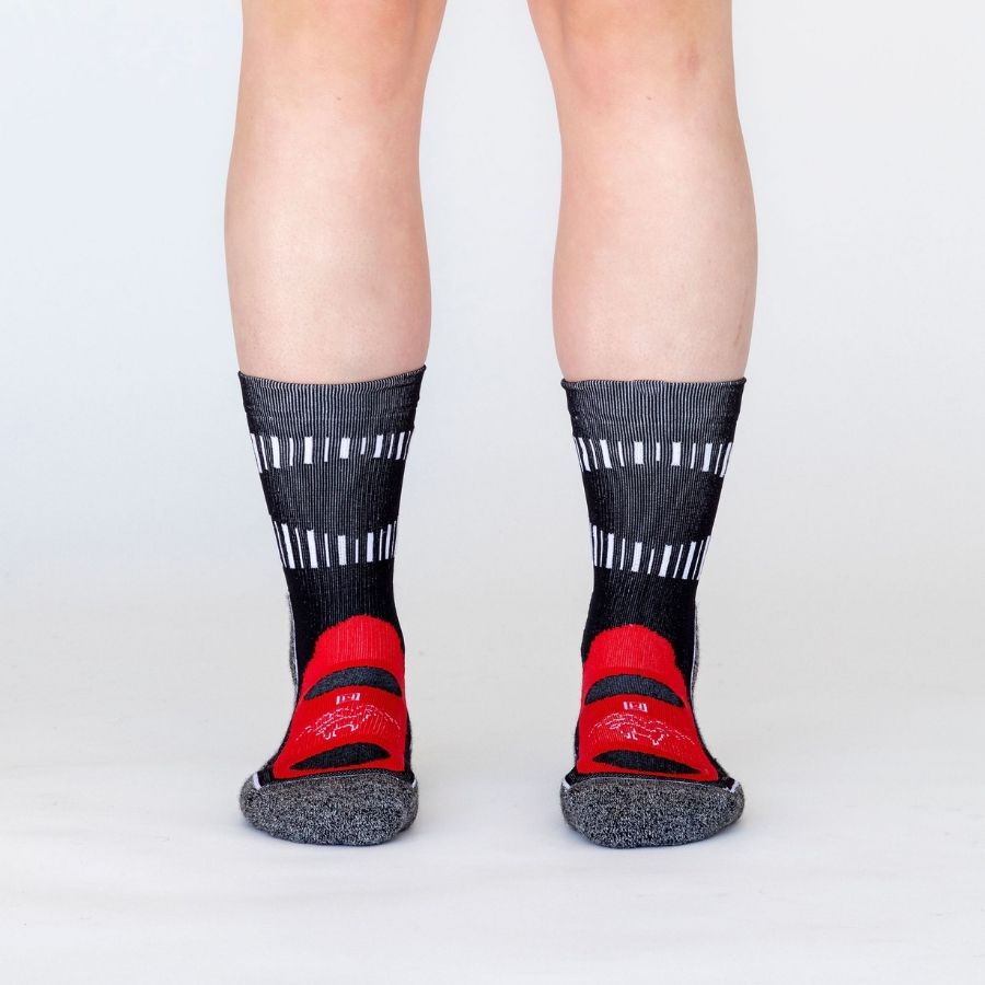 A photo of a person's lower legs standing against a white background wearing black, scarlet red, multi-gray, and white accent Alpacas of Montana soft cozy comfortable activewear outerwear athletic workout moisture wicking antimicrobial cushioned light compression engineered high-tech mid-crew hiking sock for walking, skiing, climbing, hunting, camping, fishing, exercise, biking