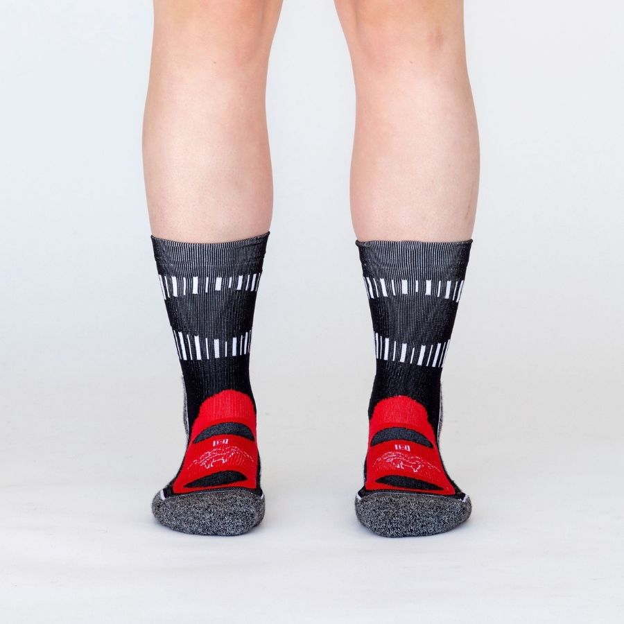 A photo of a person&#39;s lower legs standing against a white background wearing black, scarlet red, multi-gray, and white accent Alpacas of Montana soft cozy comfortable activewear outerwear athletic workout moisture wicking antimicrobial cushioned light compression engineered high-tech mid-crew hiking sock for walking, skiing, climbing, hunting, camping, fishing, exercise, biking