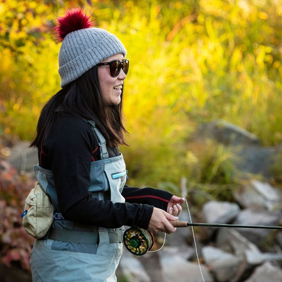 woman smiling while fly fishing and wearing a soft warm winter cozy moisture wicking comfortable fashionable light gray alpaca wool beartooth beanie with ruby red pom pom