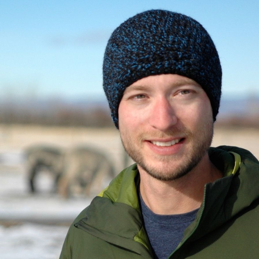 A smilling man with a brown beard standing in a sunny scene. He is wearing a green jacket and an Alpacas of Montana soft cozy comfortable stylish thermal fashionable moisture wicking antimicrobial knitted crochet mountaineer skullcap beanie hat handmade in Montana from black and bright blue alpaca wool and bamboo yarn.