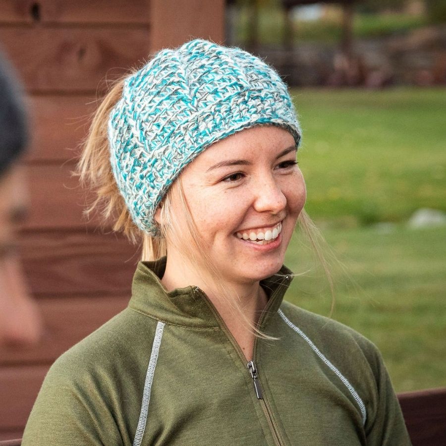 A blonde woman looking to the side and smiling. She is wearing an olive green Alpacas of Montana base layer top and a soft cozy comfortable fashionable moisture wicking knitted crochet ponytail hat handmade in Montana from natural white, bright teal turquoise blue, and multi-gray alpaca wool and bamboo yarn.