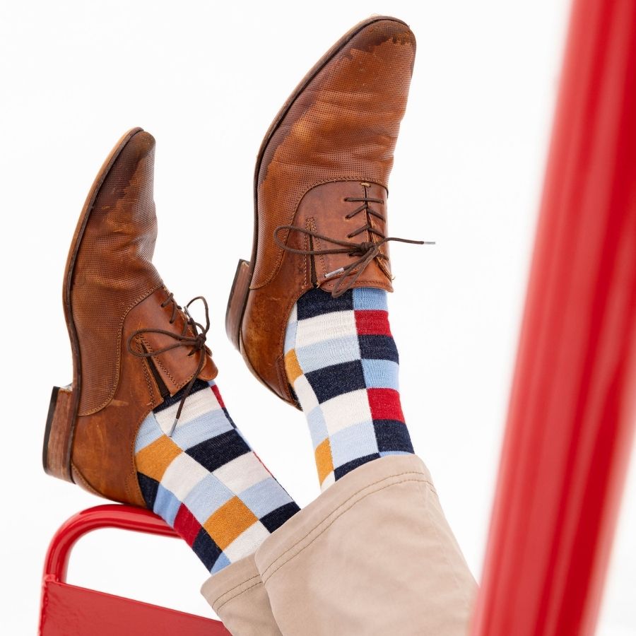 A pair of feet resting on a red chair with a white background. The feet are wearing brown dress shoes, tan pants, and Alpacas of Montana white, red, orange, navy blue, light blue, and sky blue checkered pattern casual lounge fashion comfortable soft cozy everyday moisture wicking alpaca wool Own It socks.