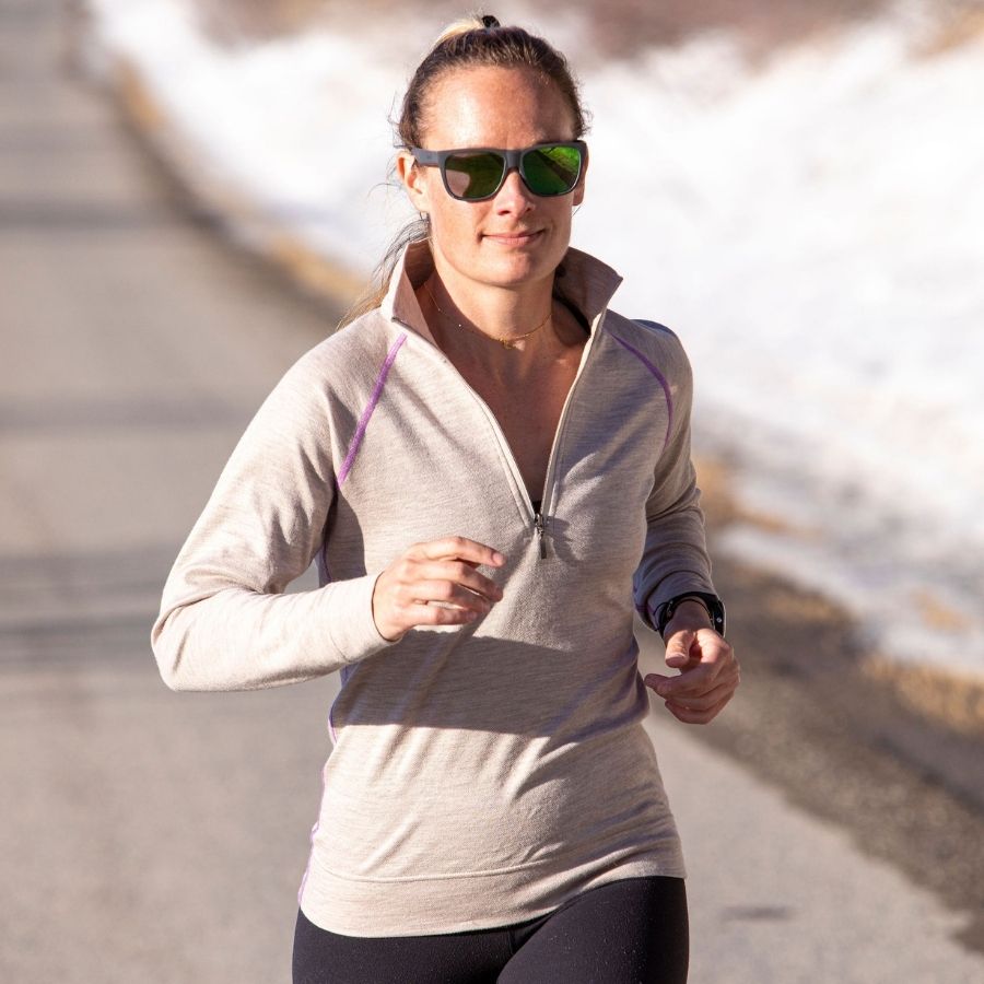 A waist-up photo of a woman running on a paved path. She is wearing back leggings and an oatmeal tan ivory with purple stitching Alpacas of Montana warm soft cozy comfortable activewear outerwear athletic moisture wicking women&#39;s fashion stylish luxury quarter-zip alpaca wool top for outdoors camping hiking skiing hunting fishing running winter.