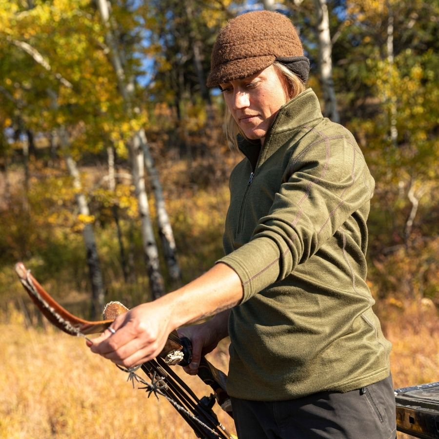 A blonde woman stringing a hunting bow in a forest scene. She is wearing an olive green Alpacas of Montana women's base layer top and a brown Alpacas of Montana an extremely warm cozy soft windproof comfortable moisture wicking thermal alpaca fleece wool windstopper winter hat for hiking, skiing, hunting, fishing, outdoors.
