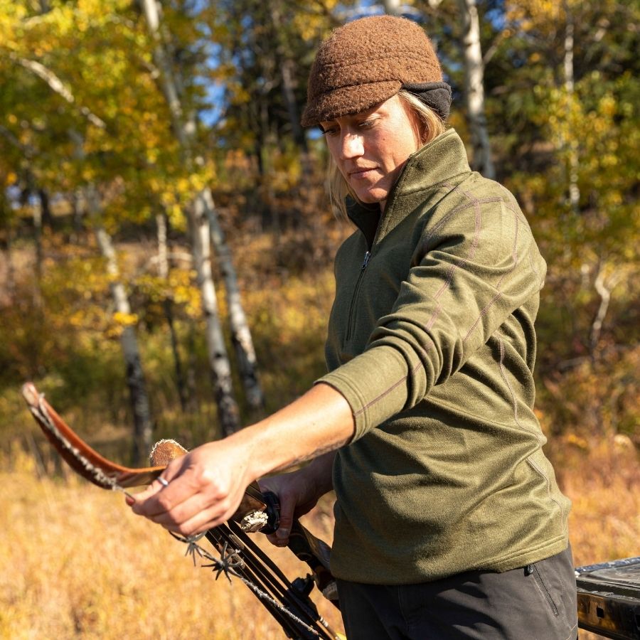 A blonde woman stringing a hunting bow in a forest scene. She is wearing an olive green Alpacas of Montana women&#39;s base layer top and a brown Alpacas of Montana an extremely warm cozy soft windproof comfortable moisture wicking thermal alpaca fleece wool windstopper winter hat for hiking, skiing, hunting, fishing, outdoors.