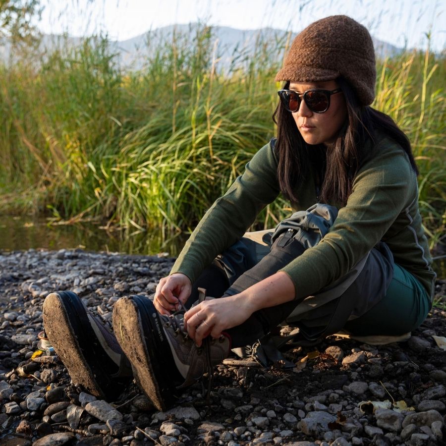 A black haired woman tying her shoe on a rocky riverbank with tall green grass in the background. She is wearing gray waders, an olive green Alpacas of Montana women&#39;s base layer top, and a brown Alpacas of Montana an extremely warm cozy soft windproof comfortable moisture wicking thermal alpaca fleece wool windstopper winter hat for hiking, skiing, hunting, fishing, outdoors.