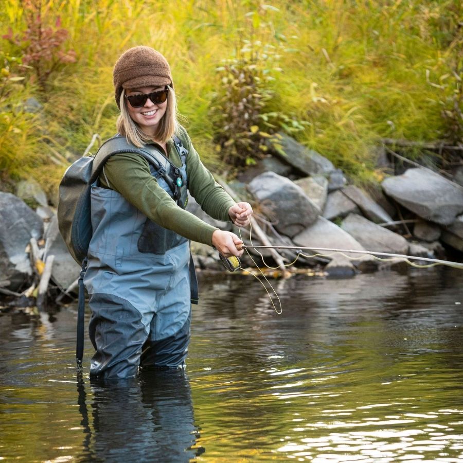 A smiling blonde woman standing and fishing in a stream with grassy banks. She is wearing light blue waders, a backpack, an Alpacas of Montana women&#39;s base layer top, and an Alpacas of Montana an extremely warm cozy soft windproof comfortable moisture wicking thermal alpaca fleece wool black windstopper winter hat for hiking, skiing, hunting, fishing, outdoors.