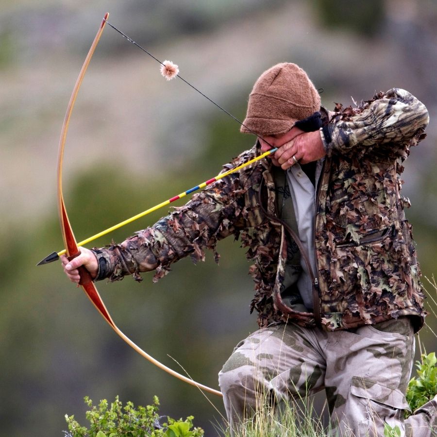 A man partially kneeling in grass and drawing back a hunting bow. He is wearing hunter&#39;s camouflage pants and jacket and a brown extremely warm cozy soft windproof comfortable moisture wicking thermal alpaca fleece wool windstopper winter hat for hiking, skiing, hunting, fishing, outdoors.