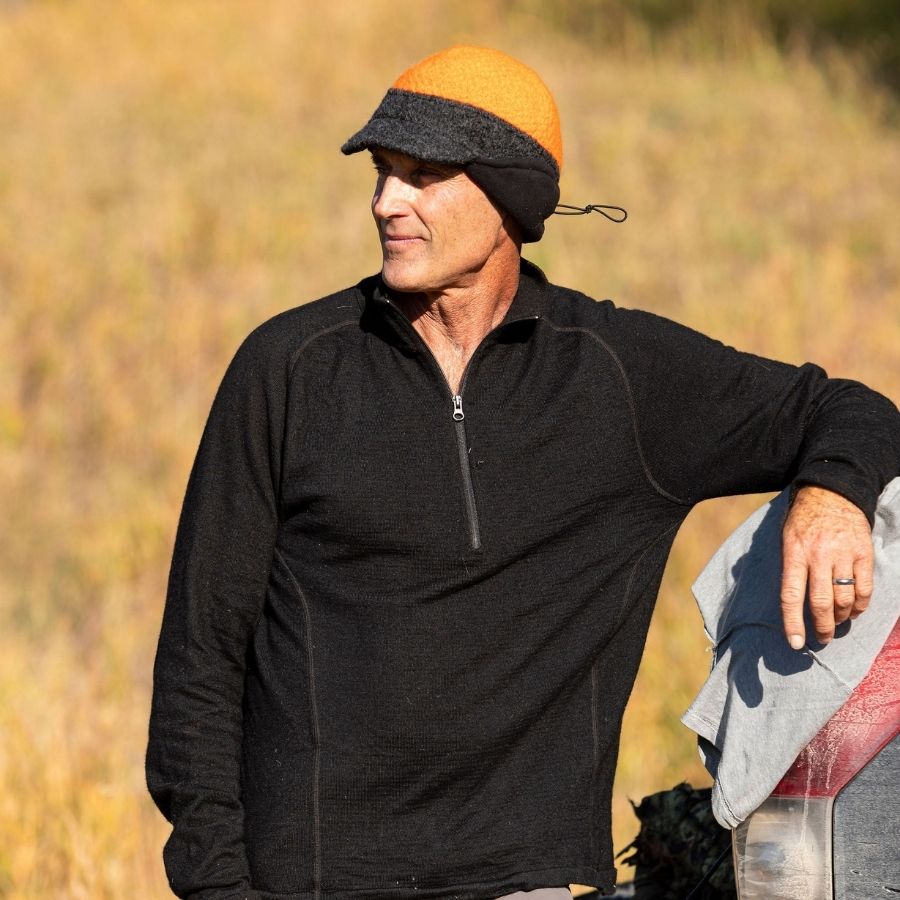 A man leaning up against the back of a truck and looking into the distance. He is wearing a black Alpacas of Montana men's mid-layer quarter zip top and an orange and gray Alpacas of Montana an extremely warm cozy soft windproof comfortable moisture wicking thermal alpaca fleece wool windstopper winter hat for hiking, skiing, hunting, fishing, outdoors.