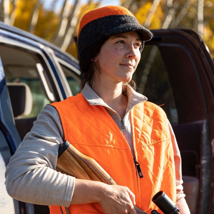 A woman standing in front of a truck's open door staring into the distance. She is wearing an orange hunting vest, an oatmeal Alpacas of Montana women's base layer top, and an orange and gray Alpacas of Montana an extremely warm cozy soft windproof comfortable moisture wicking thermal alpaca fleece wool windstopper winter hat for hiking, skiing, hunting, fishing, outdoors.