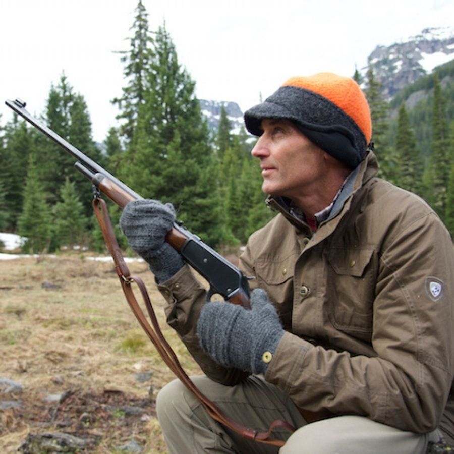 A man sitting in a forest scene. He is wearing tan pants, a brown jacket, gray mittens, and an orange and gray Alpacas of Montana an extremely warm cozy soft windproof comfortable moisture wicking thermal alpaca fleece wool windstopper winter hat for hiking, skiing, hunting, fishing, outdoors.