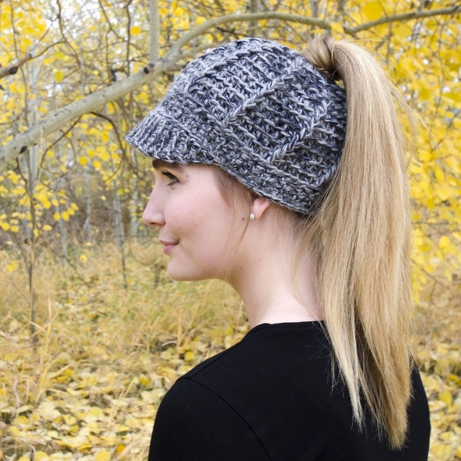 A side profile photo of a blonde woman in an autumn forest scene. She is wearing a black shirt and a soft cozy comfortable fashionable moisture wicking knitted crochet brimmed ponytail hat handmade in Montana from multi-gray and charcoal gray alpaca wool yarn.
