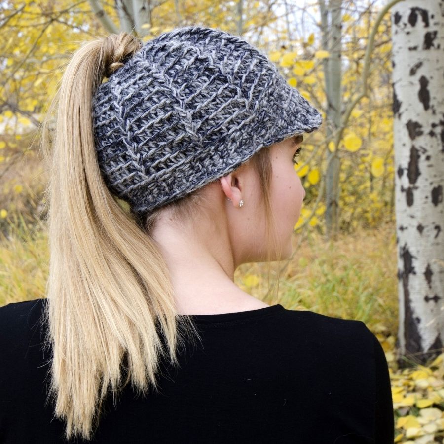 A blonde woman facing away from the camera in an autumn scene. She is wearing a black shirt and a soft cozy warm comfortable fashionable moisture wicking knitted crochet brimmed ponytail hat handmade in Montana from multi-gray and charcoal gray alpaca wool yarn.