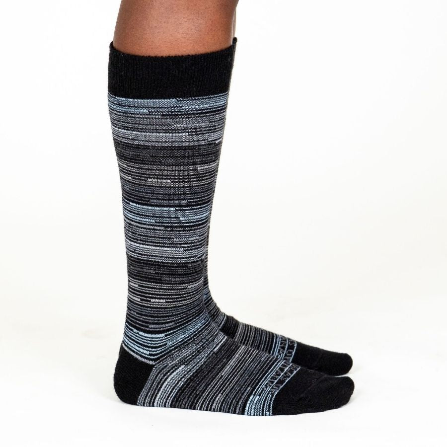 A person&#39;s lower legs standing sideways with a white background wearing Alpacas of Montana black, gray, and white casual lounge fashion comfortable soft cozy everyday moisture wicking alpaca wool striped socks.