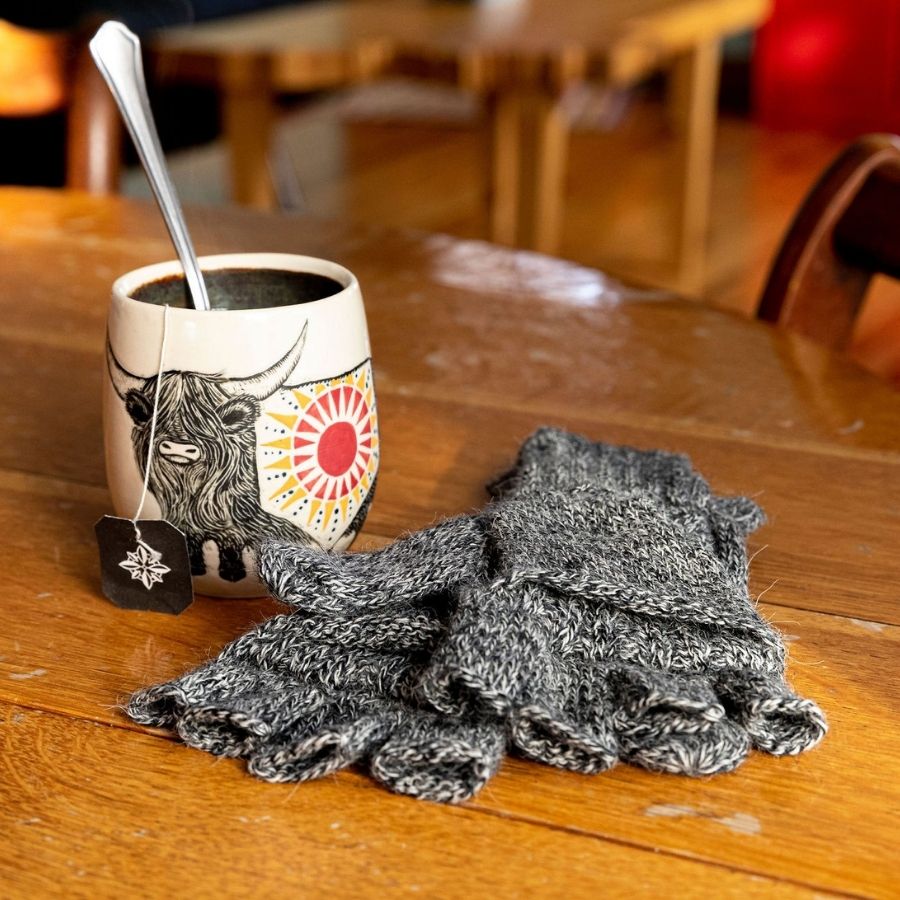 A close up photo of a white mug with a highlander cow on it and a spoon and teabag inside next to a pair of mittens on a wooden table. The mittens are the soft comfortable cozy lightweight thin cozy moisture wicking warm all seasons everyday light gray fingerless lightweight flip mitten gloves made from alpaca wool.