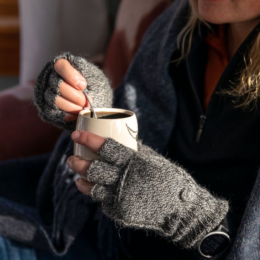 A close up of a pair of hands holding a white mug with a spoon in it. The hands are wearing soft comfortable cozy lightweight thin cozy moisture wicking warm all seasons everyday light gray fingerless lightweight flip mitten gloves made from alpaca wool.
