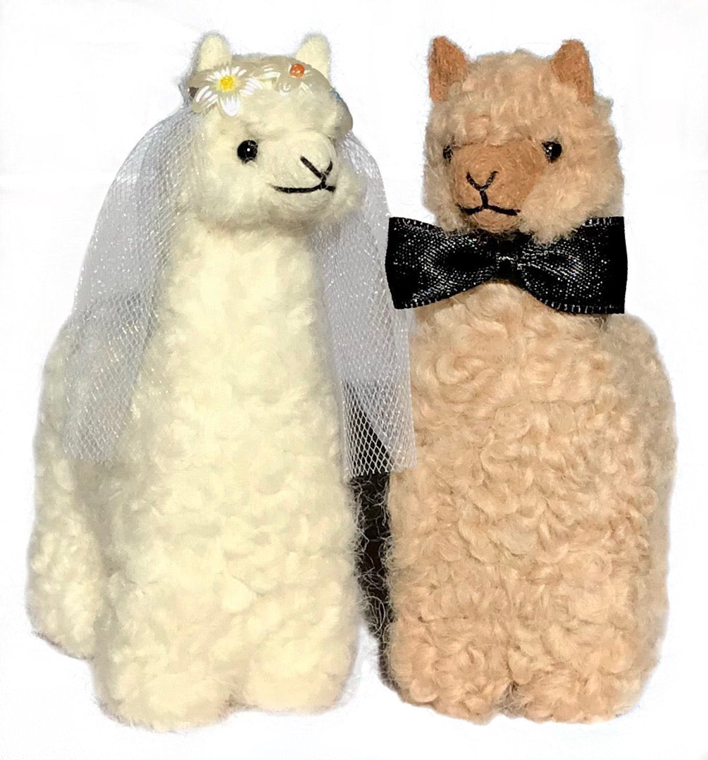 A fawn colored groom alpaca wearing a black bowtie and a white colored bride alpaca wearing a white veil and white flowers. Great for wedding gift presents wedding guests.