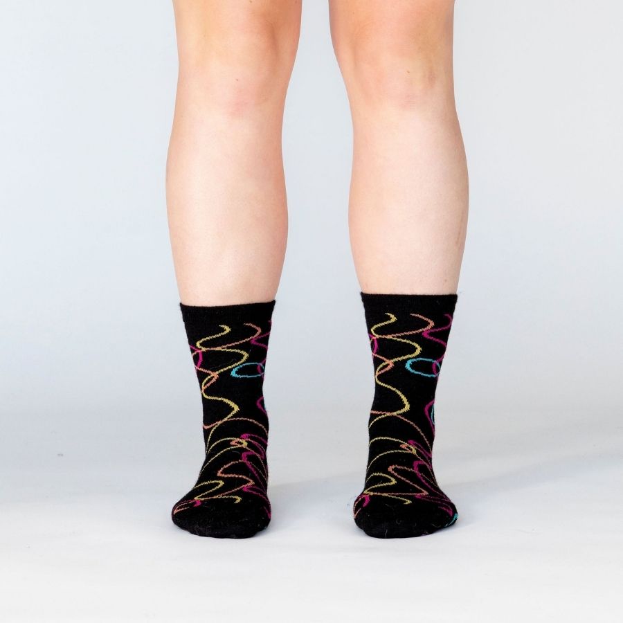 A person's lower legs against a white background wearing a pair of the Alpacas of Montana colorful yellow, orange, pink, and blue squiggle pattern black casual lounge fashion comfortable soft cozy everyday moisture wicking alpaca wool Night Life socks.