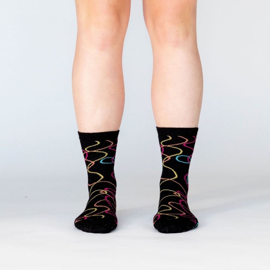 A person&#39;s lower legs against a white background wearing a pair of the Alpacas of Montana colorful yellow, orange, pink, and blue squiggle pattern black casual lounge fashion comfortable soft cozy everyday moisture wicking alpaca wool Night Life socks.
