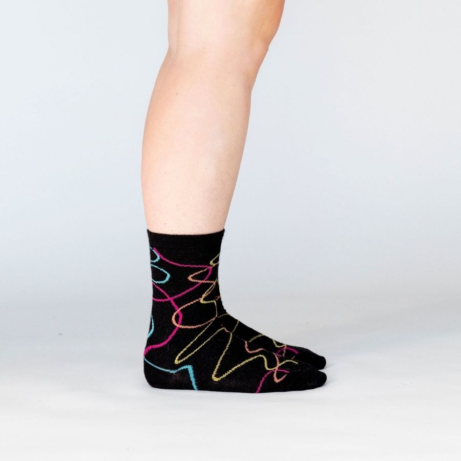 A side view of a person&#39;s lower legs with a white background wearing a pair of the Alpacas of Montana colorful yellow, orange, pink, and blue squiggle pattern black casual lounge fashion comfortable soft cozy everyday moisture wicking alpaca wool Night Life socks.