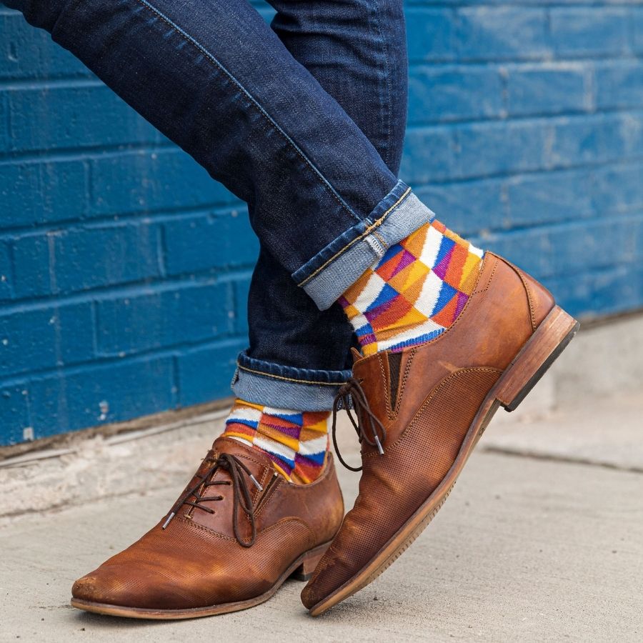A close up knees-down photo of someone standing in front of a blue brick wall. They are wearing blue jeans, chestnut brown dress shoes, and a pair of Alpacas of Montana white, yellow, gold, orange, purple, and blue triangle pattern casual lounge fashion comfortable soft cozy everyday moisture wicking alpaca wool Swag socks.