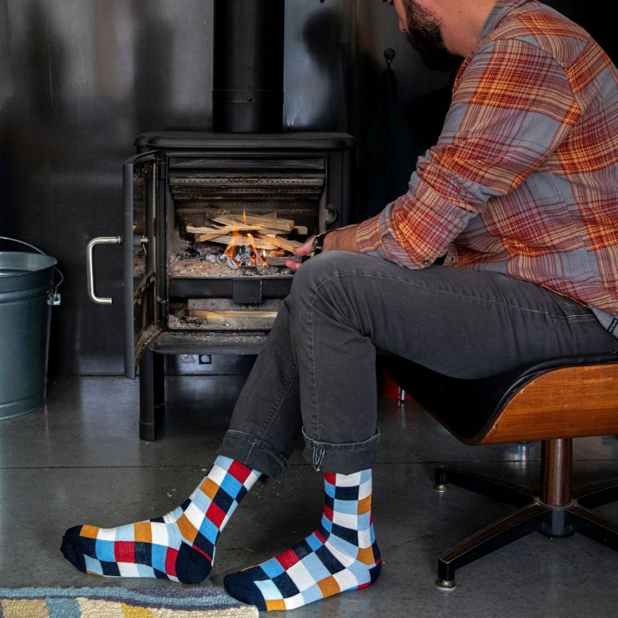 A man sitting in a wooden chair stoking a fire in a woodstove. He is wearing a red and gray plaid shirt, gray jeans, and a pair of Alpacas of Montana white, red, orange, navy blue, light blue, and sky blue checkered pattern casual lounge fashion comfortable soft cozy everyday moisture wicking alpaca wool Own It socks.