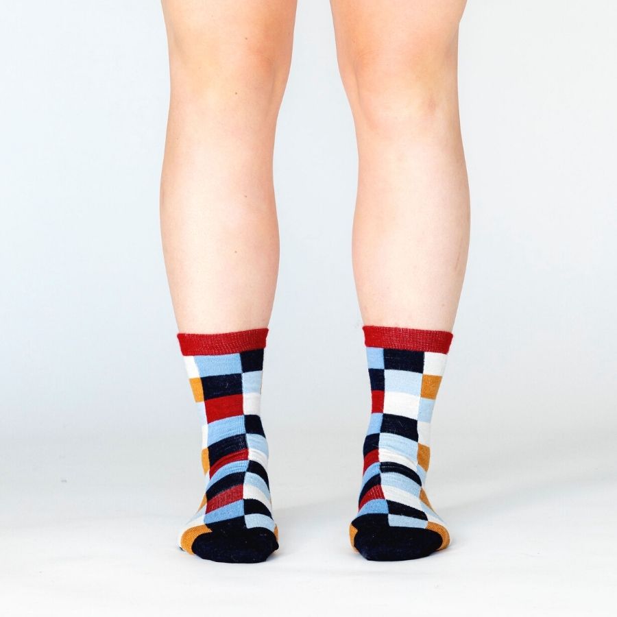 A person&#39;s lower legs standing against a white background wearing Alpacas of Montana white, red, orange, navy blue, light blue, and sky blue checkered pattern casual lounge fashion comfortable soft cozy everyday moisture wicking alpaca wool Own It socks.