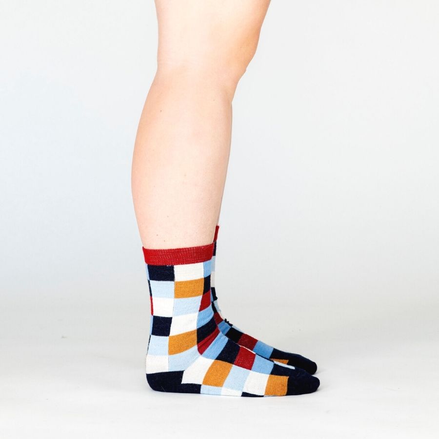 A person&#39;s lower legs standing sideways against a white background wearing a pair of Alpacas of Montana white, red, orange, navy blue, light blue, and sky blue checkered pattern casual lounge fashion comfortable soft cozy everyday moisture wicking alpaca wool Own It socks.