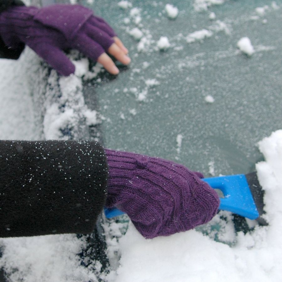A close up photo of a pair of hands using a blue tool to scrap snow off of a car windshield. The hands are wearing a pair of soft comfortable cozy lightweight thin cozy moisture wicking warm all seasons everyday purple violet fingerless lightweight flip mitten gloves made from alpaca wool.