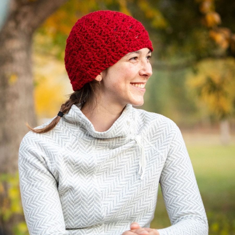 A woman with brown hair wearing a white and gray sweater and a soft stylish cozy comfortable fashionable moisture wicking knitted crochet scallop pattern hat handmade in Montana from scarlet red alpaca wool.