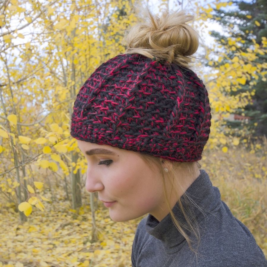 A blonde woman standing in an autumn scene wearing a gray shirt and a soft cozy comfortable fashionable moisture wicking knitted crochet ponytail hat handmade in Montana from black and red alpaca wool and bamboo yarn.