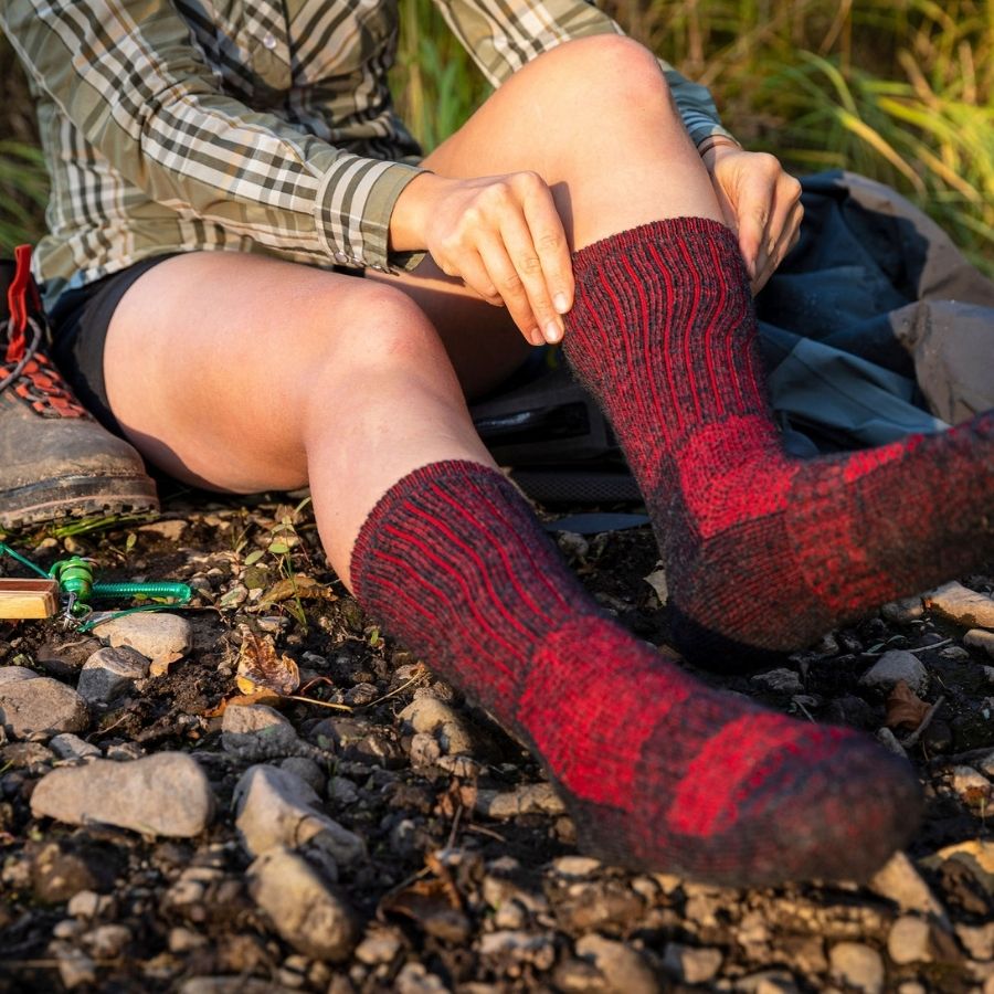 A person sitting outdoors wearing a plaid shirt and black shorts. They are pulling on a pair of Alpacas of Montana cozy soft warm comfortable thermal moisture wicking everyday winter fishing hiking snowshoeing hunting outdoors scarlet and black wine red extra cushion boot socks.