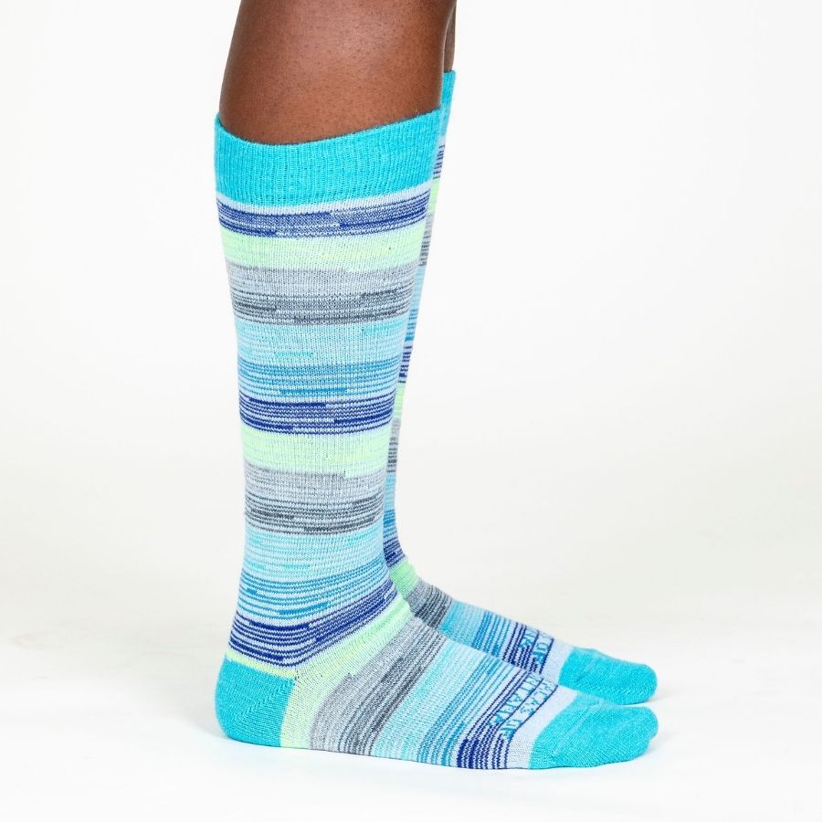 A person&#39;s lower legs standing sideways with a white background wearing Alpacas of Montana colorful navy, cobalt, sky blue, lime green, gray, and black casual lounge fashion comfortable soft cozy everyday moisture wicking alpaca wool striped socks.