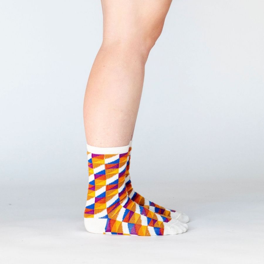 A person&#39;s lower legs standing sideways wearing a pair of Alpacas of Montana white, yellow, gold, orange, purple, and blue triangle pattern casual lounge fashion comfortable soft cozy everyday moisture wicking alpaca wool Swag socks.